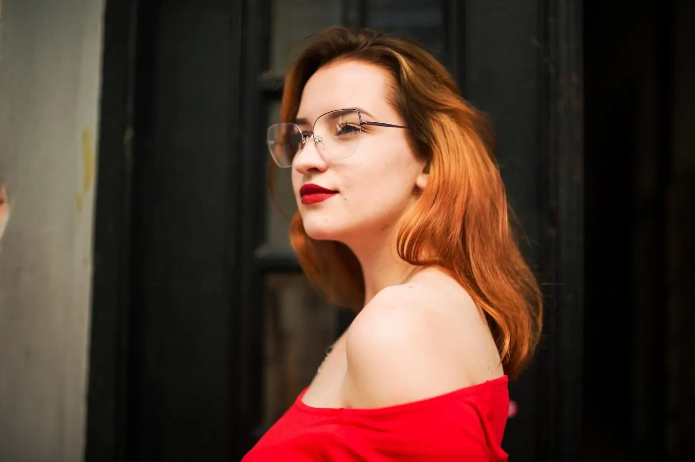 An attractive red-haired woman in eyeglasses, wearing a red blouse, posing in the street against an old wooden door.
